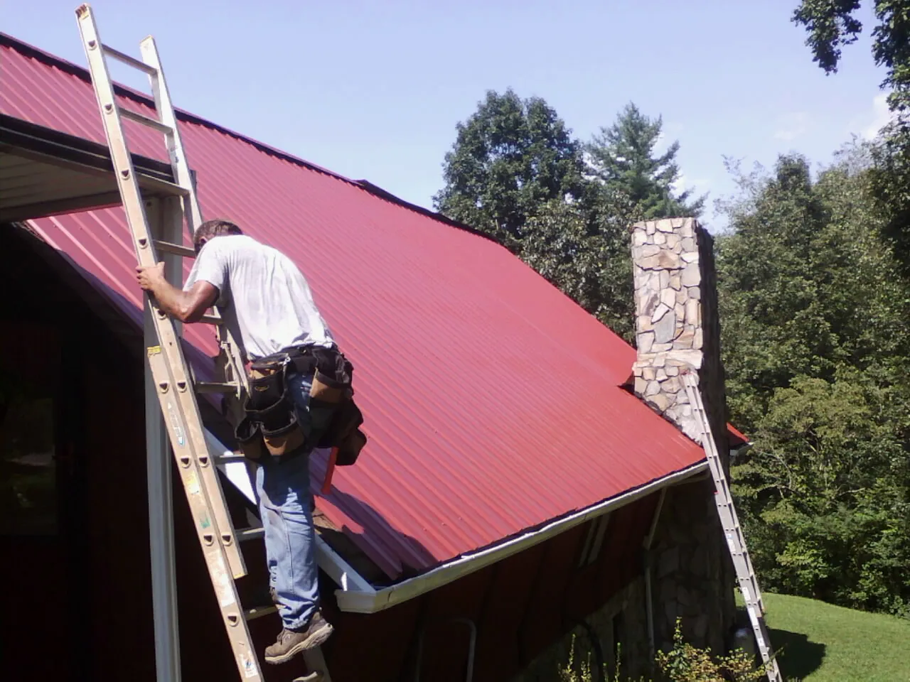 Roofer on a ladder beside a red metal roof and stone chimney — Soffit & Fascia Repair by Roofing Brothers in North Franklin